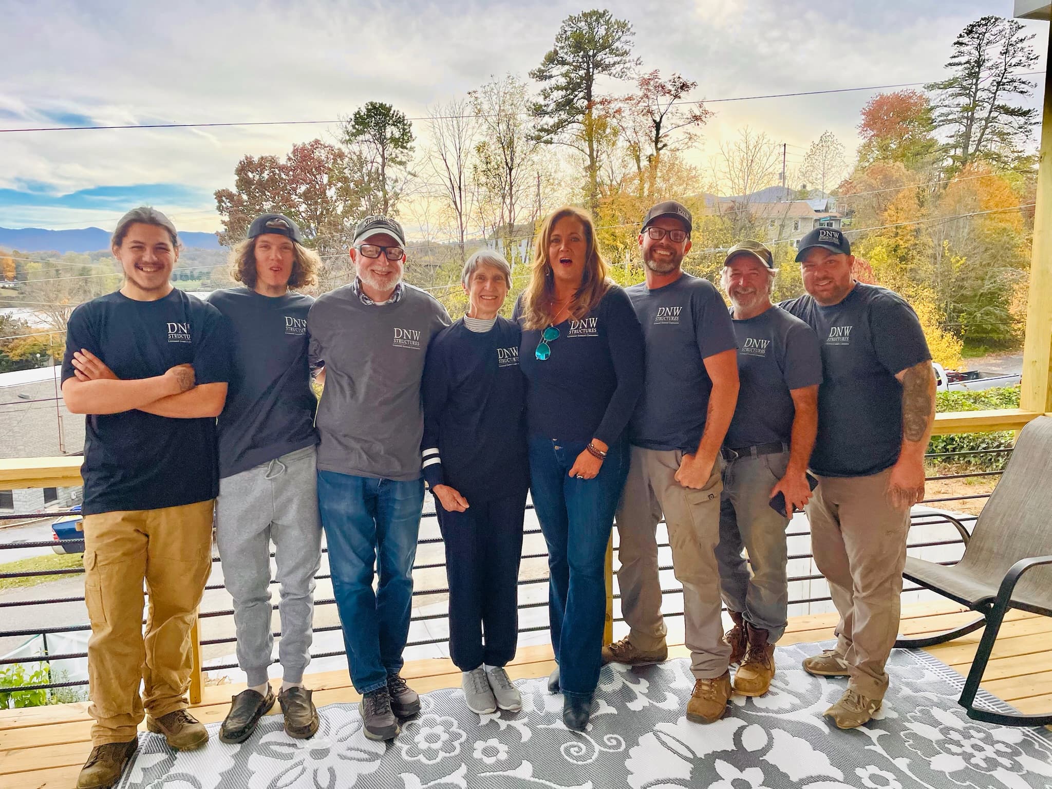 Eight people in DNW Structures shirts pose on a wooden deck with scenic mountain background.