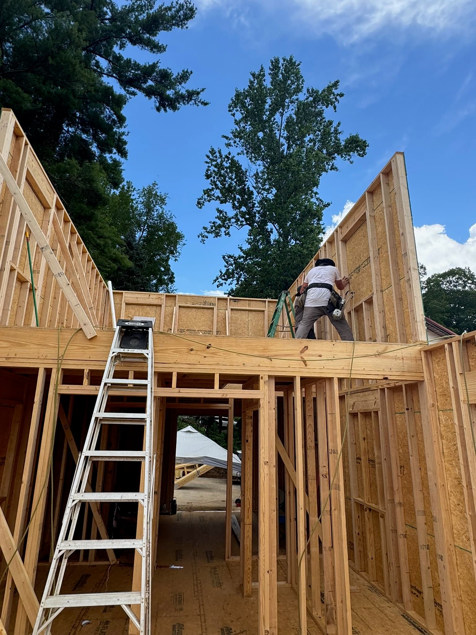 Construction worker framing a wooden house on a sunny day with a ladder in foreground.